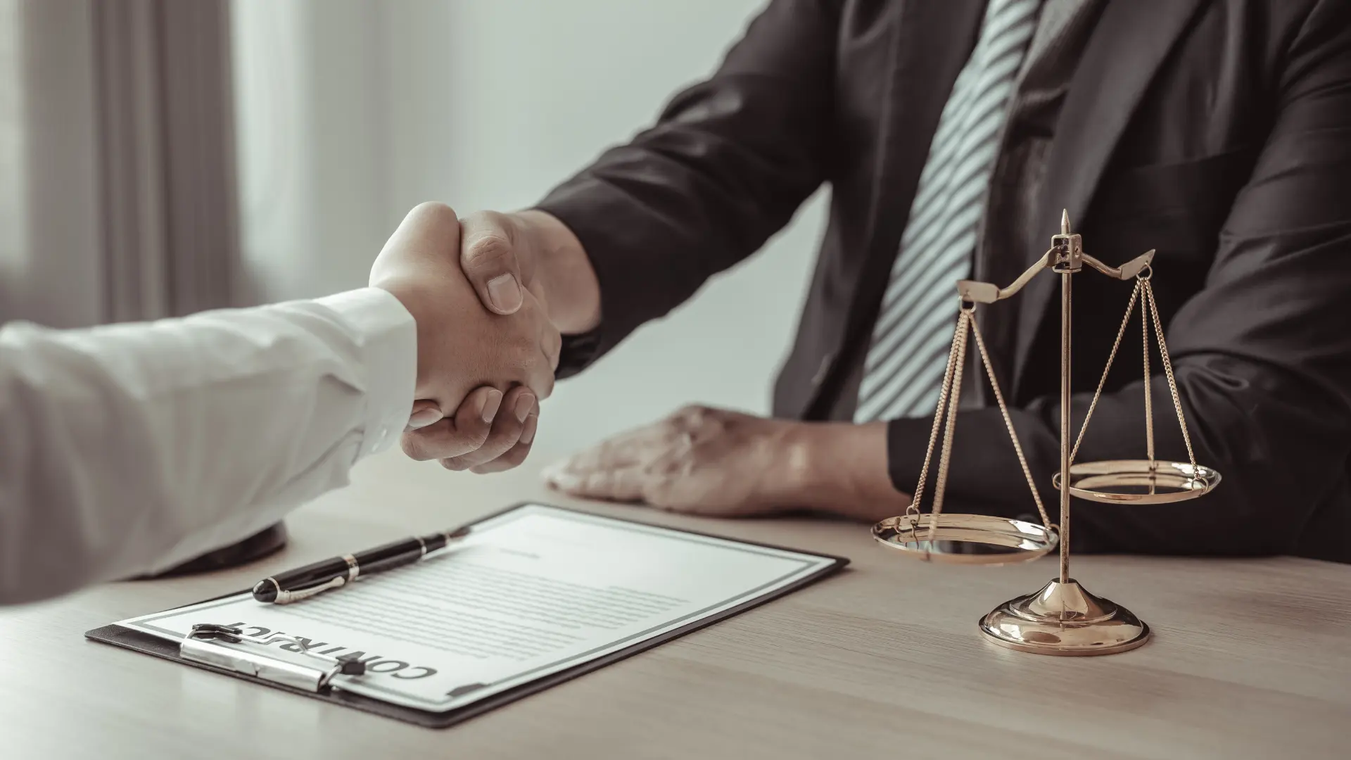 Two people shaking hands over legal documents with scales of justice on the table.